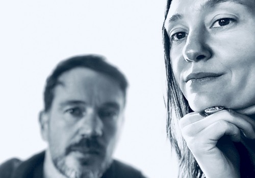 Two people pose for a close-up black and white photo, with one woman in focus and a man blurred in the background.