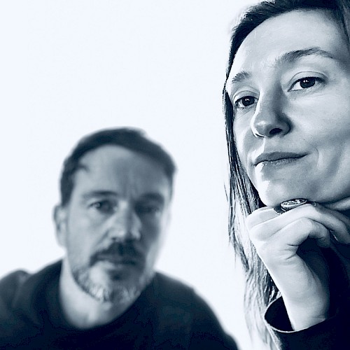 Two people pose for a close-up black and white photo, with one woman in focus and a man blurred in the background.
