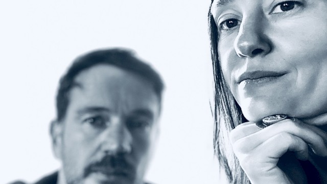 Two people pose for a close-up black and white photo, with one woman in focus and a man blurred in the background.