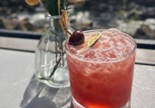 A pink cocktail with a cherry, lemon slice, and decorative greenery in a glass on a sunny windowsill.