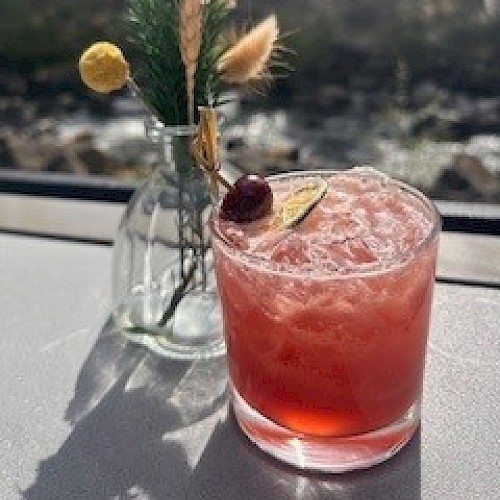 A pink cocktail with a cherry, lemon slice, and decorative greenery in a glass on a sunny windowsill.