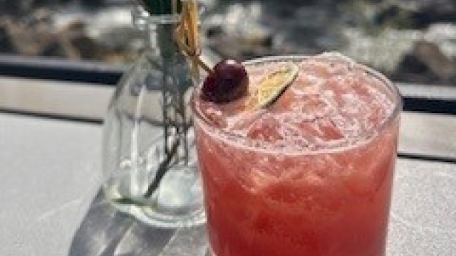 A pink cocktail with a cherry, lemon slice, and decorative greenery in a glass on a sunny windowsill.