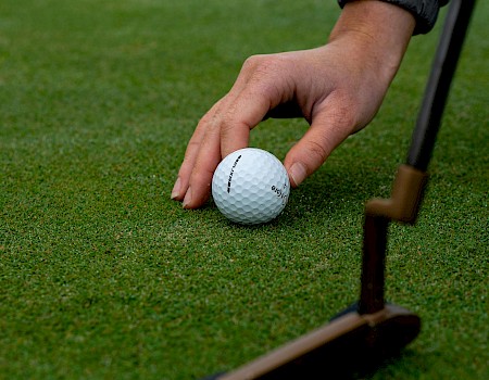 A person prepares to putt with a golf ball on a green, holding the ball, close to a golf club, on a well-maintained golf course surface.