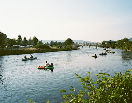 People are kayaking and paddleboarding on a river under a clear sky with greenery on the banks.