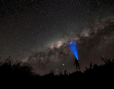 A person with a flashlight under a starry night sky, pointing towards the Milky Way galaxy in a silhouette view.
