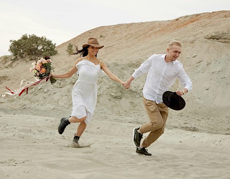 A couple is running hand-in-hand across a sandy desert landscape, the woman holding a bouquet and both smiling happily.