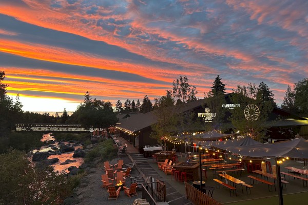 Riverhouse Lodge in Bend, Oregon is shown at sunset, with string lights, tables, chairs, trees, a bridge, and colorful sky.