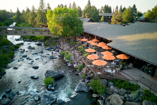 This image features Currents restaurant with orange umbrellas along a rocky riverbank at Riverhouse Lodge in Bend Oregon, surrounded by green trees and residential buildings.