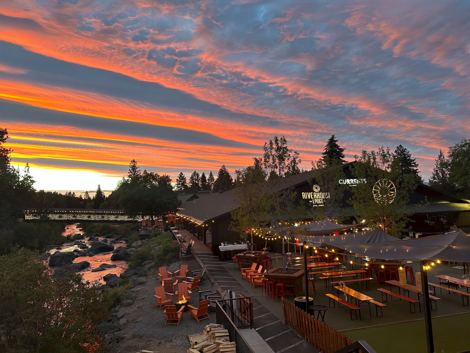 Riverhouse Lodge in Bend, Oregon at sunset with outdoor seating, string lights, trees, a bridge, and a dramatic colorful sky.