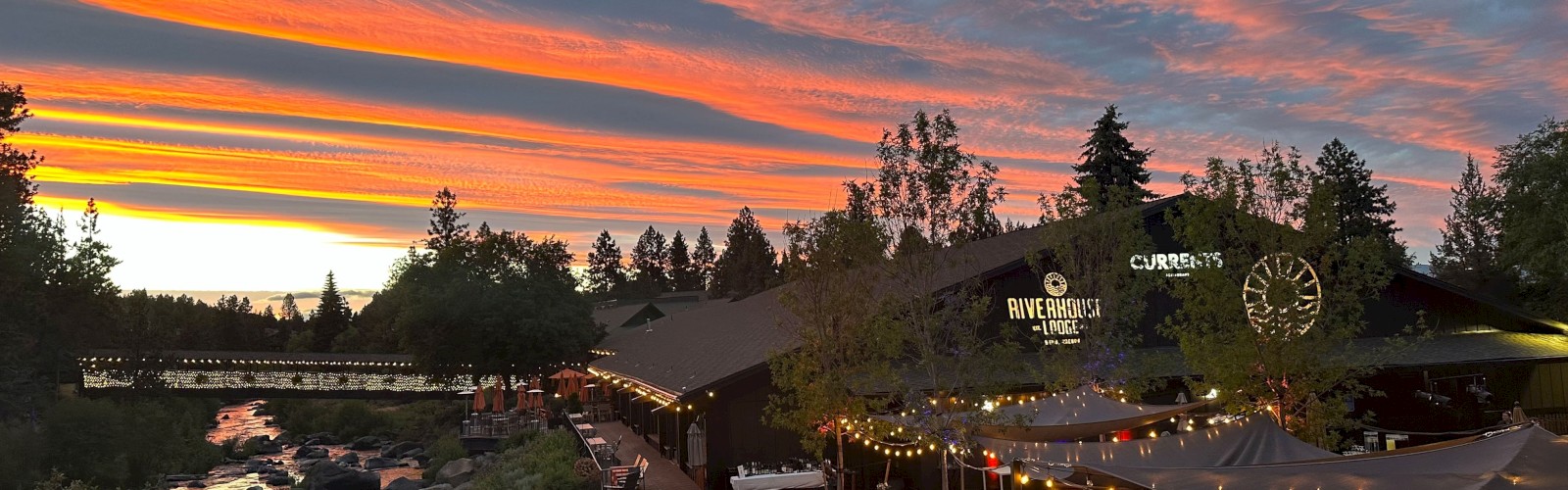 Riverhouse Lodge in Bend, Oregon at sunset with outdoor seating, string lights, trees, a bridge, and a dramatic colorful sky.