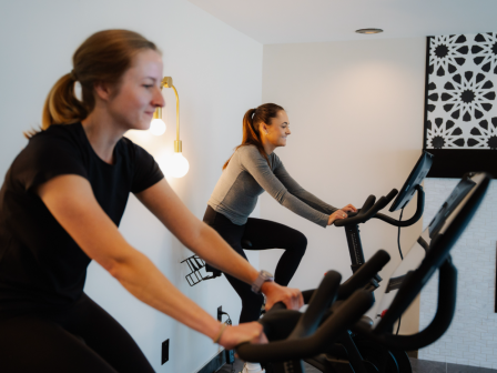 Two women are exercising on stationary bikes indoors, smiling and enjoying their workout session.