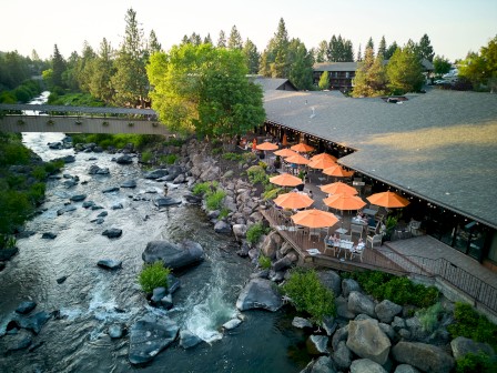 A riverside outdoor dining area with orange umbrellas, next to a rocky stream surrounded by trees and greenery.