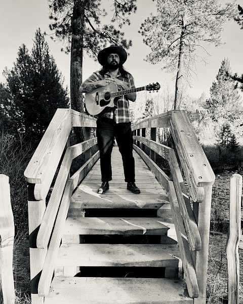 Dom Ruben stands on a wooden bridge holding an acoustic guitar among Central Oregon pine trees, promoting live music performances at Currents Restaurant & Lounge at Riverhouse Lodge in Bend, Oregon.