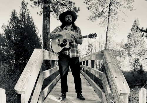 Dom Ruben stands on a wooden bridge holding an acoustic guitar among Central Oregon pine trees, promoting live music performances at Currents Restaurant & Lounge at Riverhouse Lodge in Bend, Oregon.