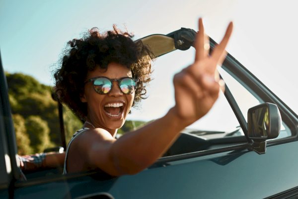 A woman with curly hair and sunglasses is smiling and making a peace sign while leaning out of a car window on a sunny day.