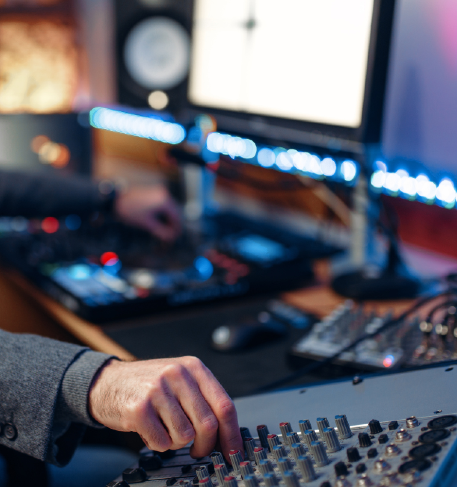 A person with a beard in a gray blazer DJing with audio equipment and monitors in a cozy, lit studio.