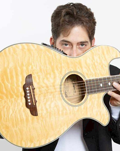 A man holds an acoustic guitar partially hiding his face, looking directly at the camera, against a plain, light background.