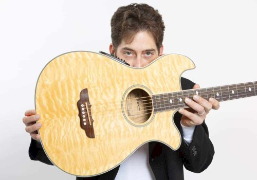 A man holds an acoustic guitar partially hiding his face, looking directly at the camera, against a plain, light background.