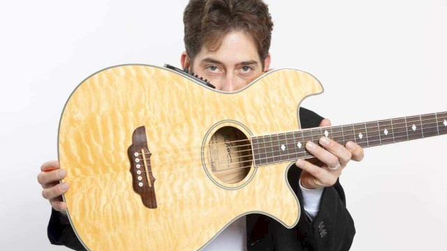 A man holds an acoustic guitar partially hiding his face, looking directly at the camera, against a plain, light background.