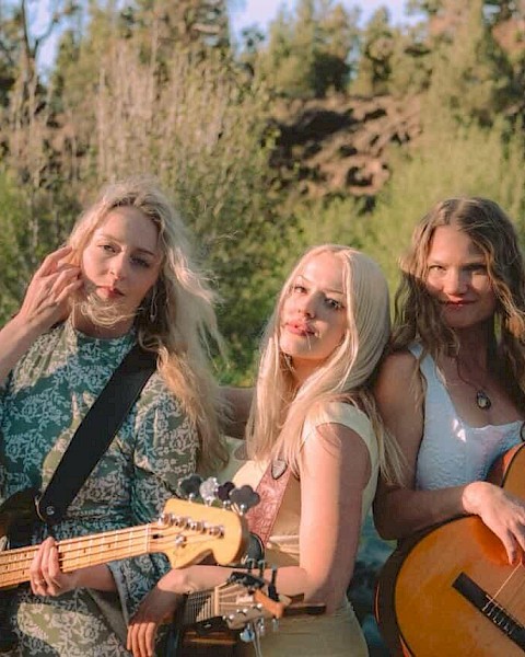 Three women with guitars pose by a river with rocks and greenery, enjoying a sunny day outdoors.