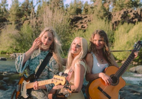 Three women with guitars pose by a river with rocks and greenery, enjoying a sunny day outdoors.