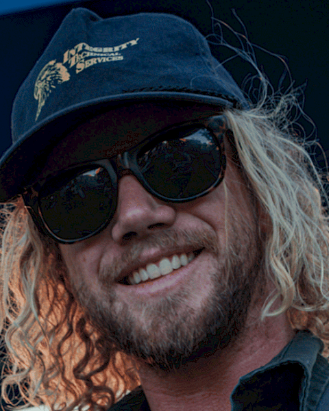 A man with curly blonde hair, sunglasses, a cap, and a beard is smiling outdoors.