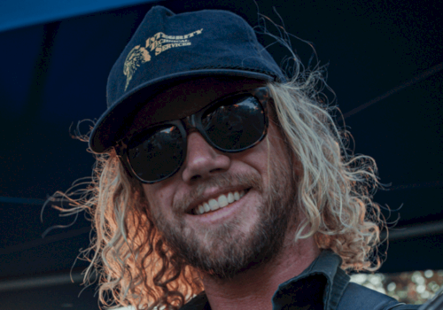 A man with curly blonde hair, sunglasses, a cap, and a beard is smiling outdoors.