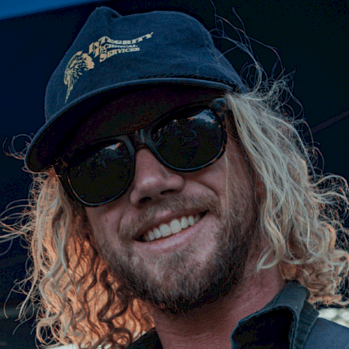 A man with curly blonde hair, sunglasses, a cap, and a beard is smiling outdoors.
