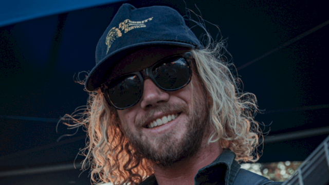 A man with curly blonde hair, sunglasses, a cap, and a beard is smiling outdoors.