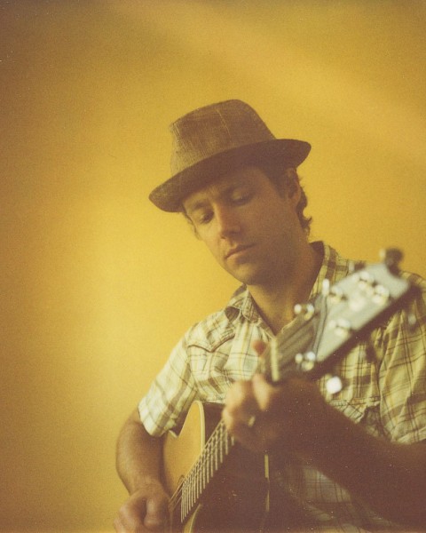 A man wearing a hat is playing an acoustic guitar against a yellow wall.