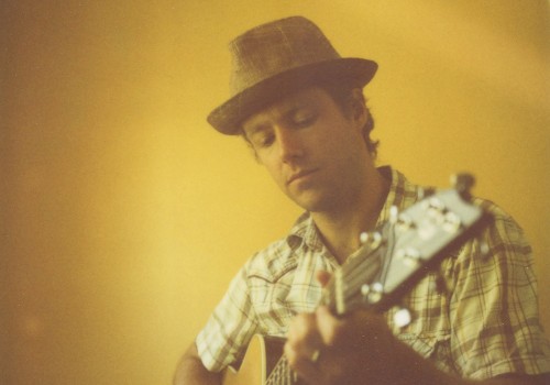 A man wearing a hat is playing an acoustic guitar against a yellow wall.