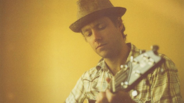 A man wearing a hat is playing an acoustic guitar against a yellow wall.