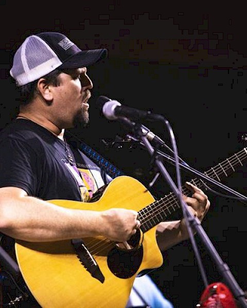 A man wearing a trucker hat is playing an acoustic guitar and singing into a microphone on stage.