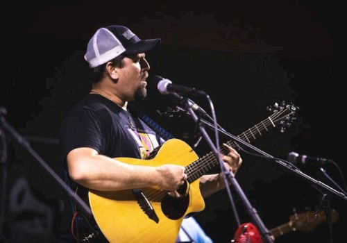 A man wearing a trucker hat is playing an acoustic guitar and singing into a microphone on stage.
