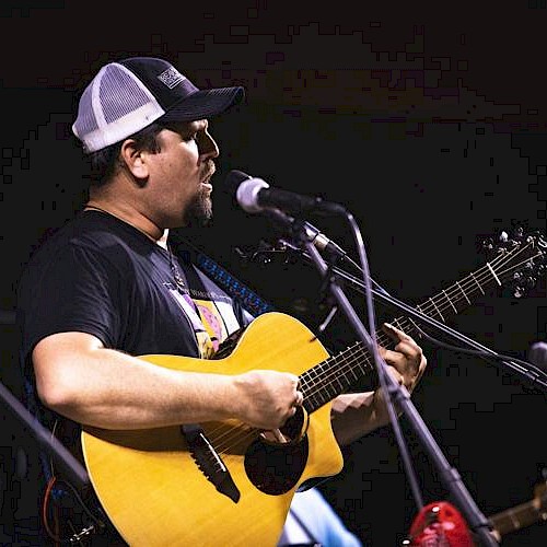 A man wearing a trucker hat is playing an acoustic guitar and singing into a microphone on stage.