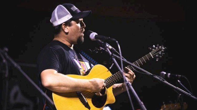 A man wearing a trucker hat is playing an acoustic guitar and singing into a microphone on stage.