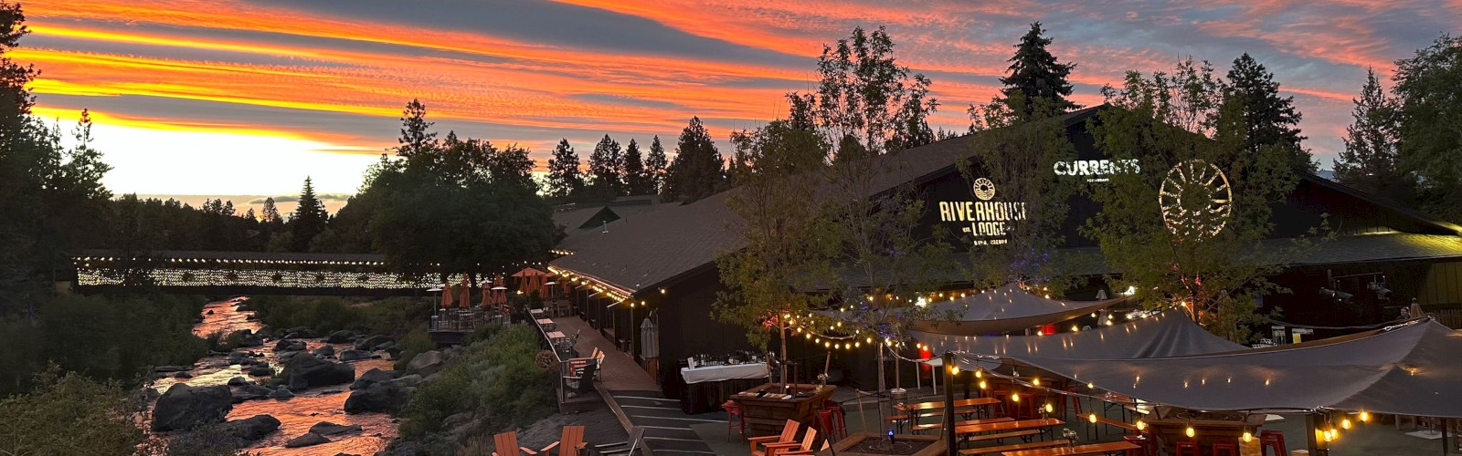 A scenic sunset over riverside dining, with string lights, outdoor tables, and a bridge in a forested area.