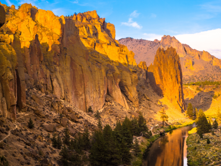 A scenic view of a canyon with towering rocky cliffs, a winding river, and lush trees under a blue sky, bathed in golden sunlight.