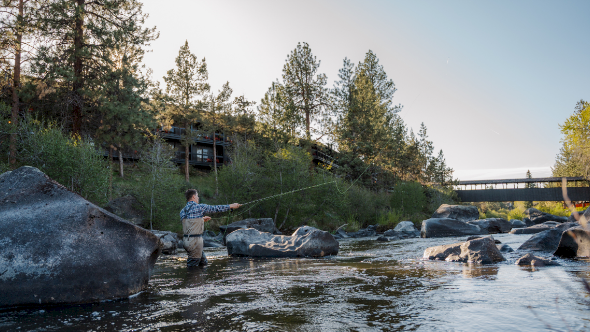A man fly fishing in a river surrounded by rocks, trees, and a bridge in a peaceful outdoor setting during daylight.
