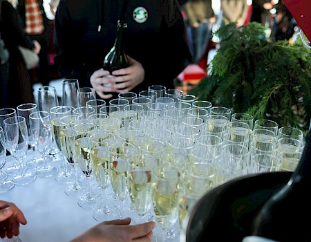 People are serving and drinking champagne at a celebration or event, with glasses arranged on a white table and others holding bottles.