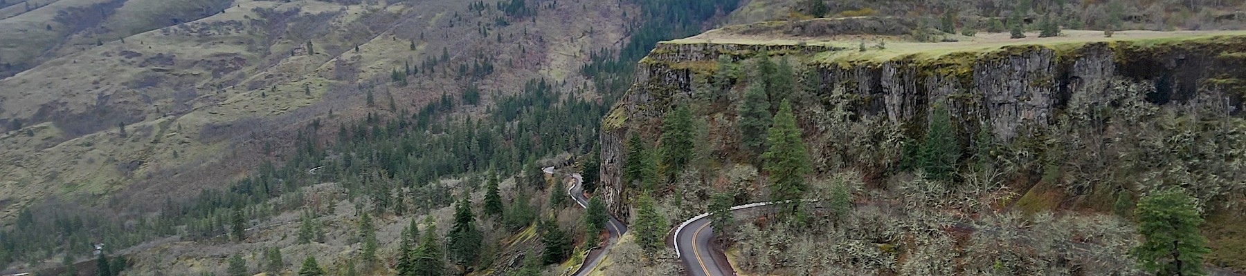 A winding mountain road with sharp curves goes through a forested hillside under a cloudy sky.