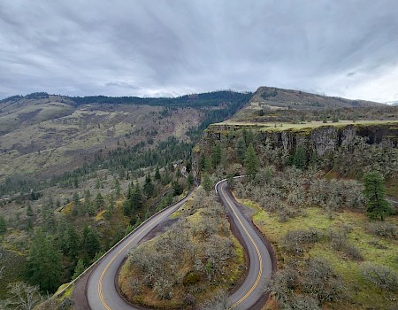 A winding mountain road with sharp curves goes through a forested hillside under a cloudy sky.