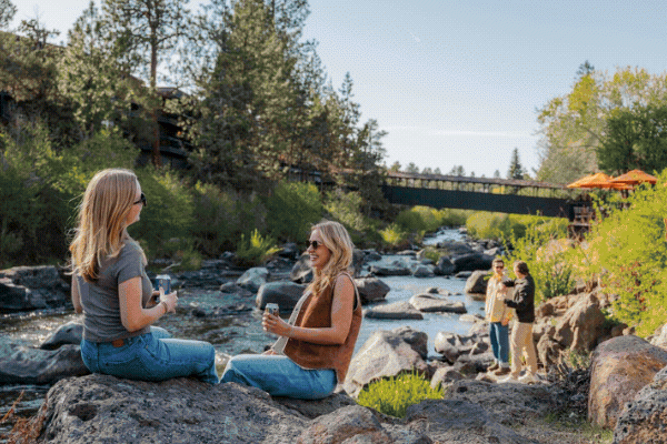 People enjoy a sunny day by a river, sitting on rocks, talking, and taking photos amidst trees and a bridge in the background.
