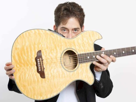 A person holds an acoustic guitar in front of their face, with only eyes visible, against a plain background.