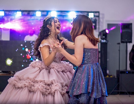 Two girls in elegant dresses are dancing and laughing at a celebration, with colorful lights and a DJ in the background.
