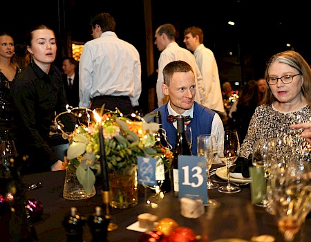 People sit and stand around a decorated table at a festive dinner event, with a mix of attentive, talking, and standing individuals.