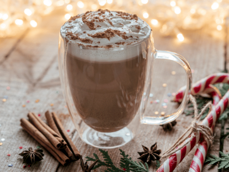 A festive hot chocolate topped with whipped cream and cocoa powder, surrounded by candy canes, cinnamon sticks, star anise, and holiday lights.