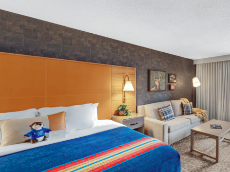 This is a modern hotel room with a bed, sofa, wall art, and a balcony overlooking greenery and trees.