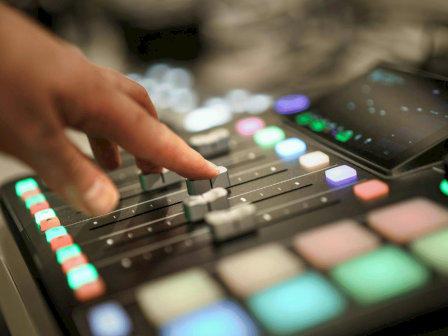 A hand is adjusting controls on a digital music mixer with colorful buttons and a screen, used for audio mixing and sound production.