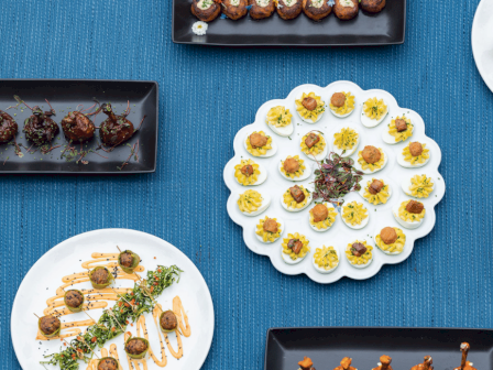 Various gourmet appetizers are displayed on black and white plates on a textured blue tablecloth, including deviled eggs, meatballs, and skewers.